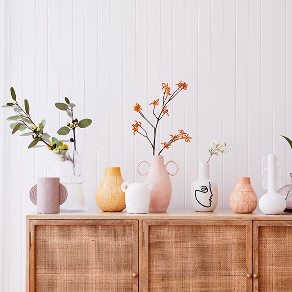 Selection of colourful vases with native floral stems sitting in a handful of the vases, all on top of a rattan sideboard.