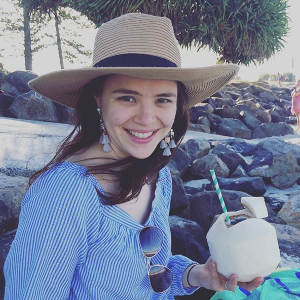 A woman smiling sitting on a beach with a coconut drink in hand and sunhat. 