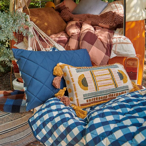 The back of a campervan is opened, with a hammock strung in front filled with cushions and a blue gingham blanket. The campervan is filled with blankets and cushions, and a knitted ottoman is visible in the left of the frame.