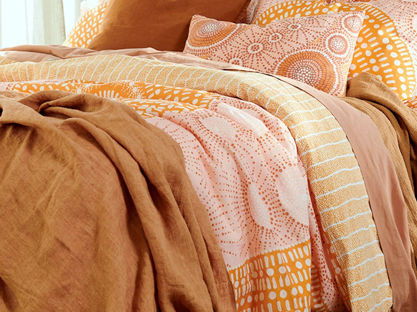 A styled bed in a focused shot displays the layered quilt cover in lighter shades of yellow, pink and orange while a rust linen throw and solid earth coloured sheets provide additional dimension. 