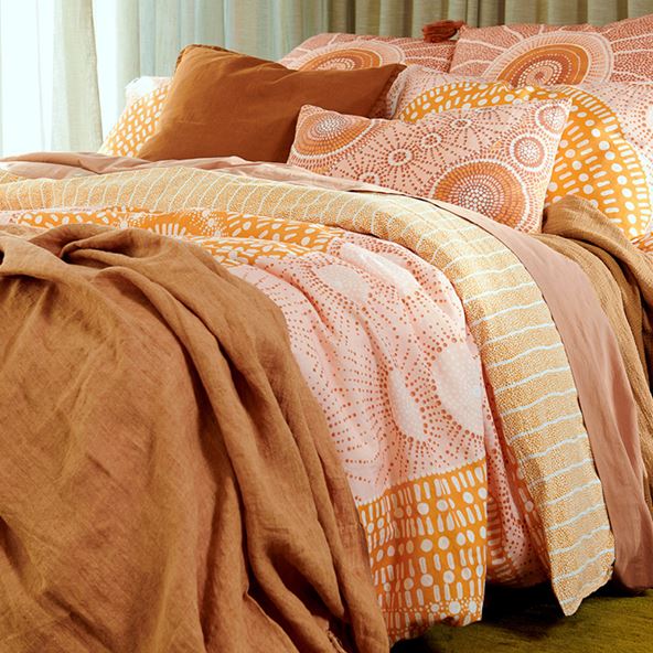 A styled bed in a focused shot displays the layered quilt cover in lighter shades of yellow, pink and orange while a rust linen throw and solid earth coloured sheets provide additional dimension. 