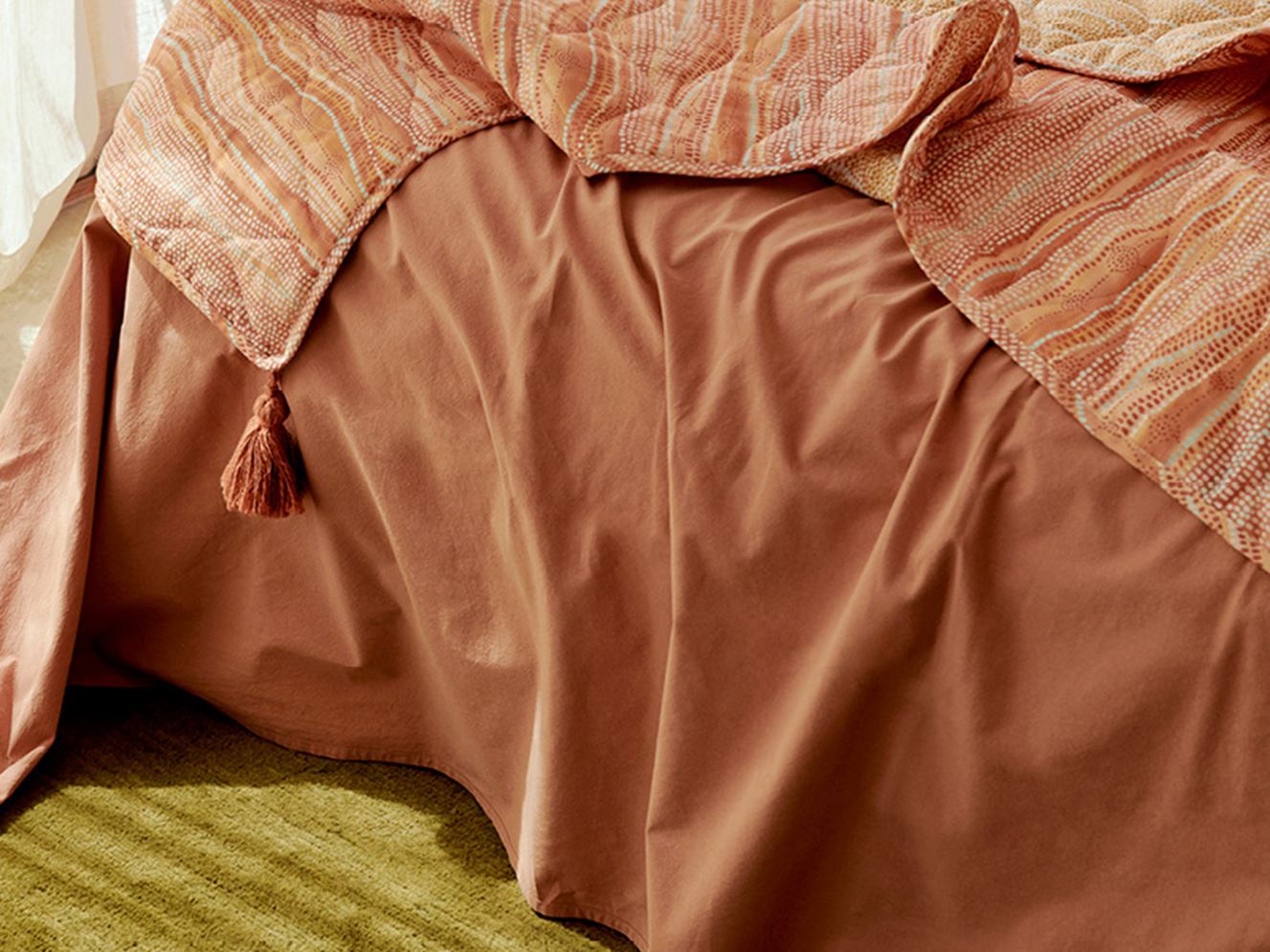 Solid earth coloured sheets styled on a bed, with a coverlet draped naturally over the top in contrasting lighter hues of orange, pink and yellow.