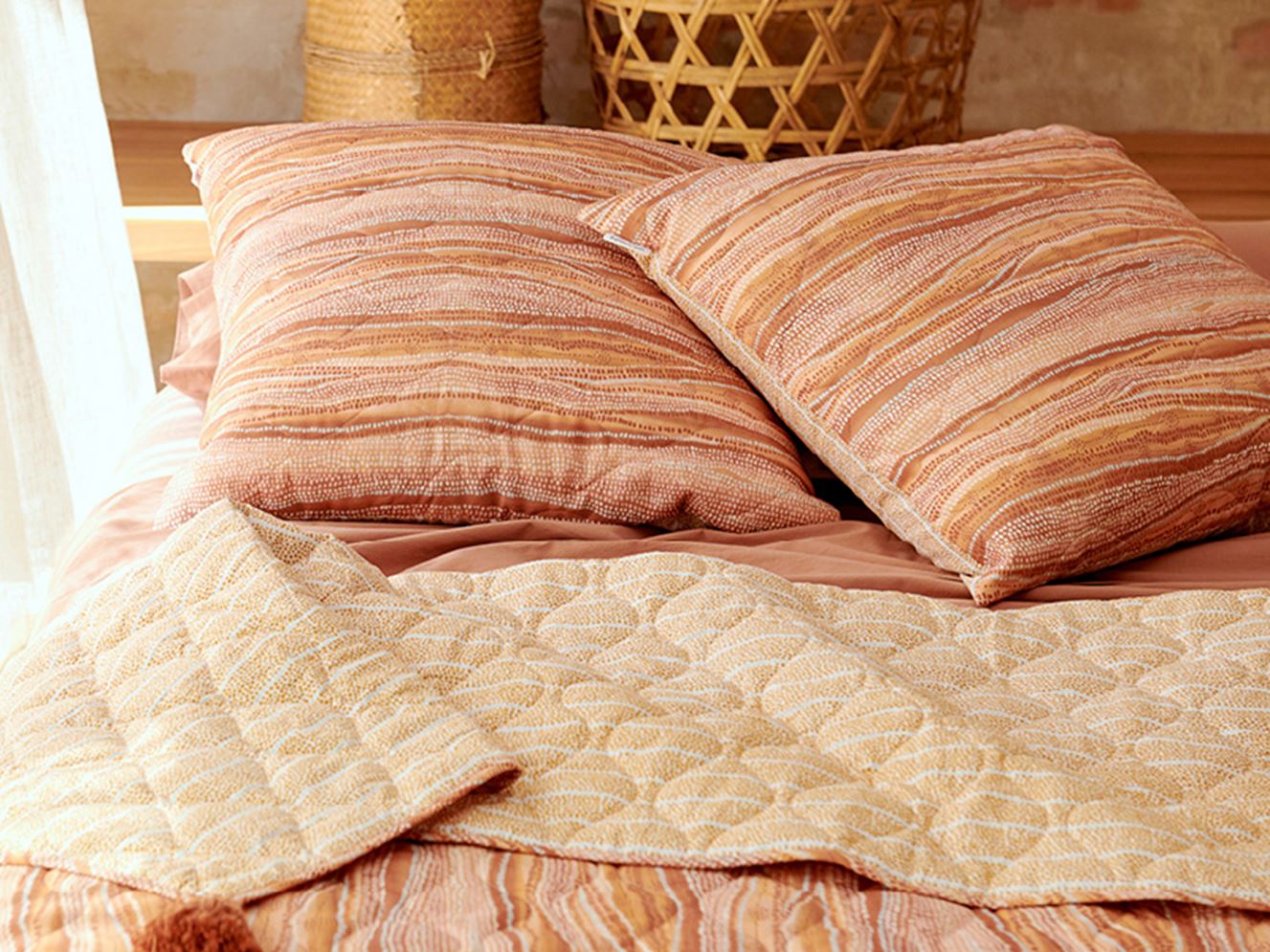 Two cushions styled on a bed to accompany the coverlet, in tones of earth and lighter shades of pink, yellow and orange.