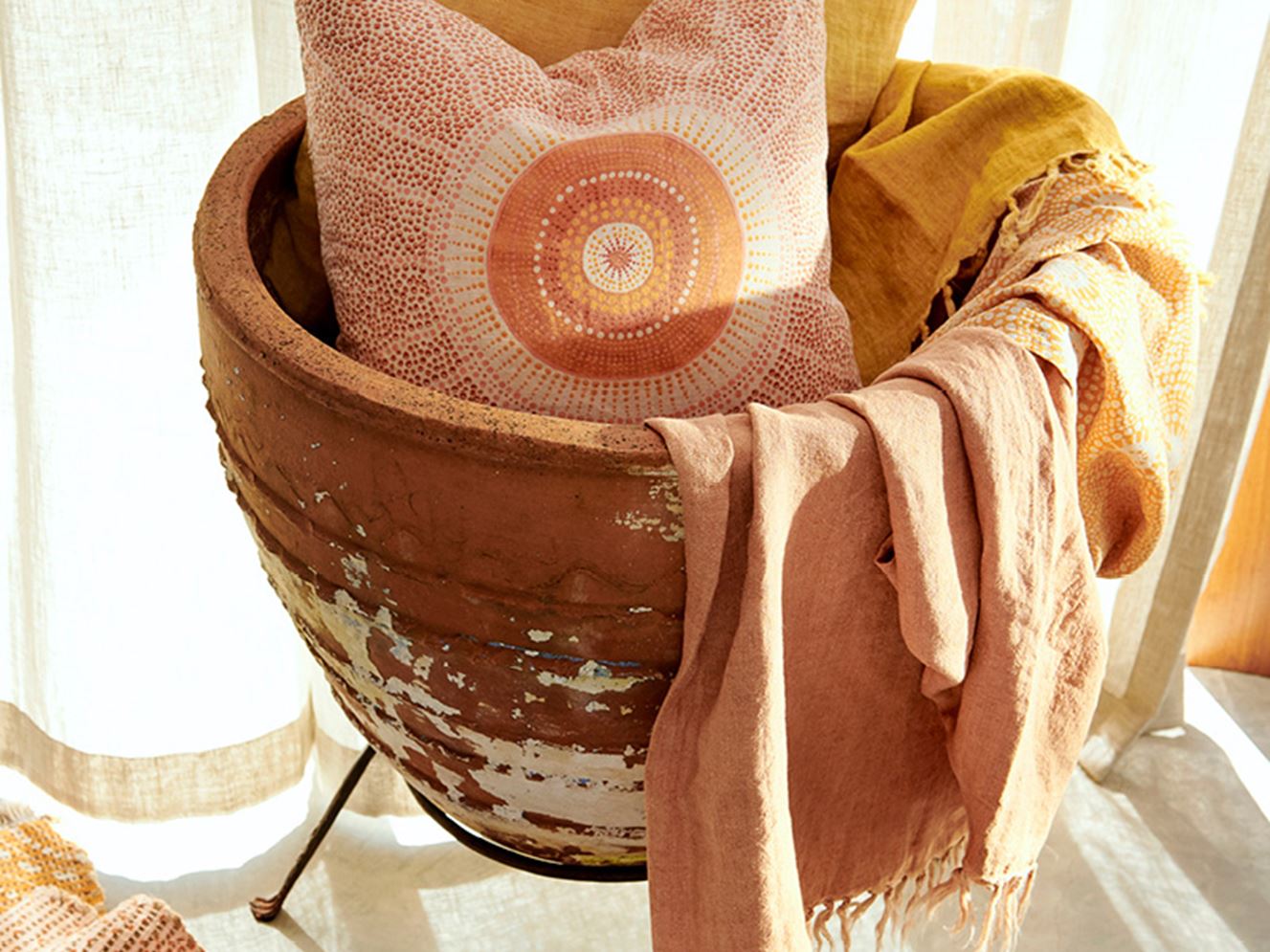 A rustic ceramic basket holds a cushion from the collection and draped linen throws, cushion has dotted design in red with bolder orange tones in centre.