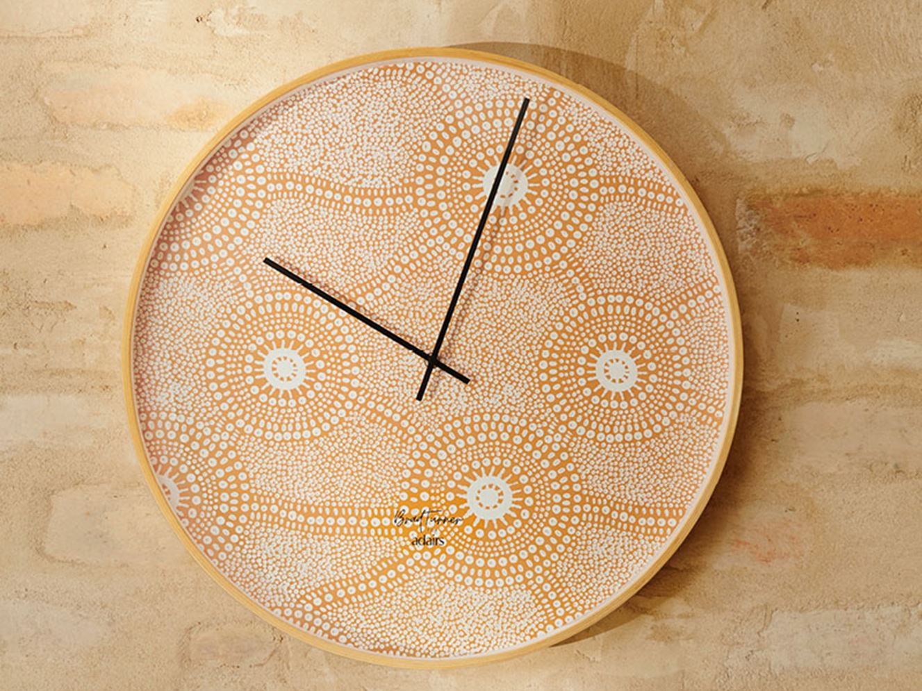 Circular analog wall clock against an earthy toned brick wall, with defined black hands contrasting against the vibrant orange dotted design. 