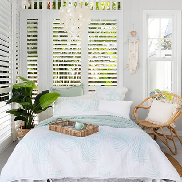 A front-on image of the coastal inspired bedroom with light blue and white bedlinen, rattan chair, tray, and pot plant.