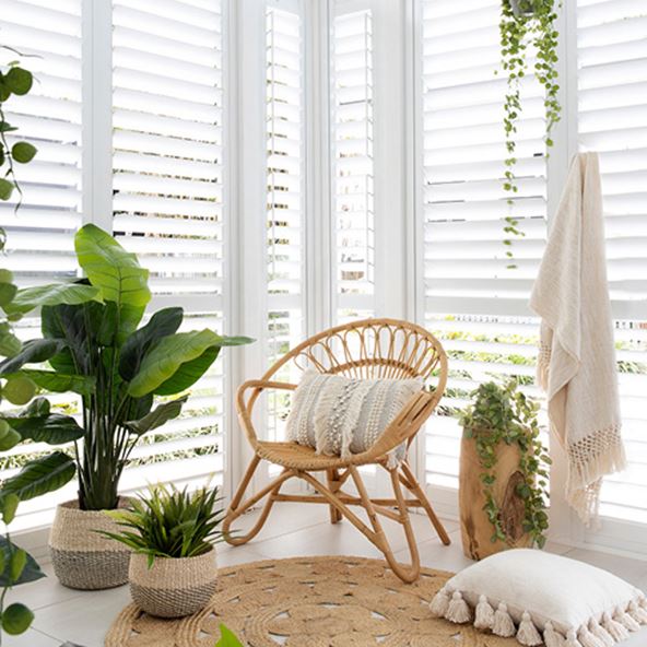 The corner of a room featuring white shutters, styled with a rattan chair, natural accessories and a multitude of plants. 