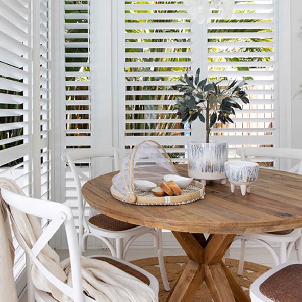 Corner of a room with white shutters, featuring a dining table with provincial chairs and styled with natural accessories. 