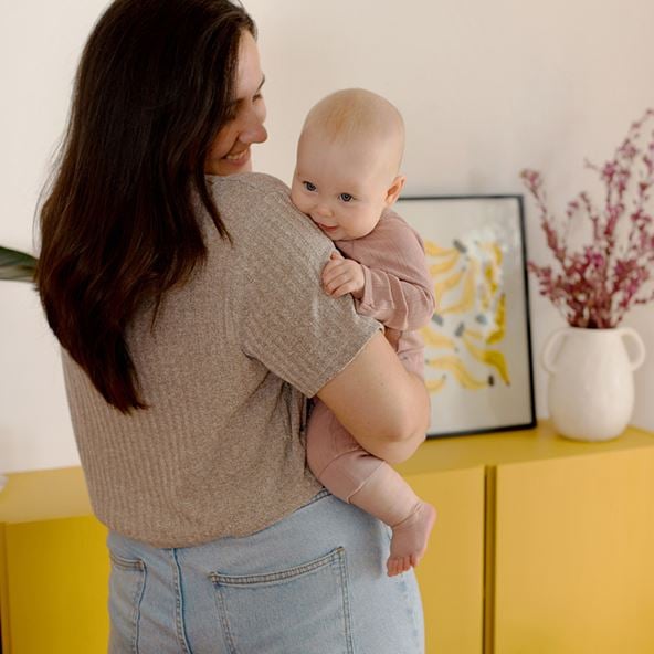 Claudia stands holding her baby with her back to the camera. Behind her is a yellow sideboard with plants and art on top.
