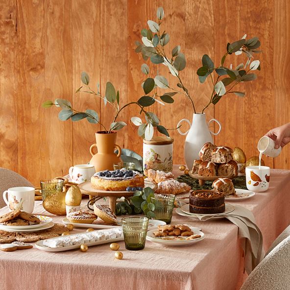 A dining room scene with a pink tablecloth and lots of baked Easter treats along the centre, served on assorted platters. Eucalyptus stems in sculptural vases are in the middle of the table, and the background is a wooden wall.