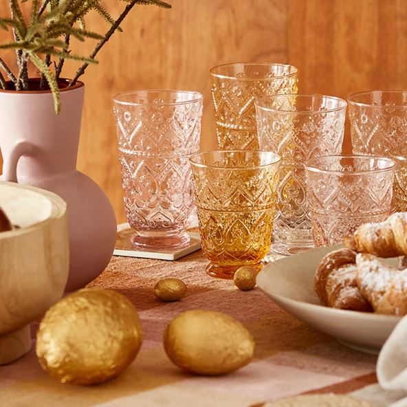 Assorted glasses in clear and orange colours, with intricate details set on coasters and a gingham tablecloth. To the sides of the frame are a plate of croissants at the left and a vase of stems to the right. There are golden easter eggs on the table.