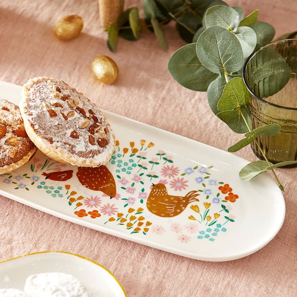 A close up and cropped image of three platters containing baked treats, the centre platter is long and features a design on chickens and flowers. In the top of the frame, slightly out of focus is a eucalyptus stem and a green glass.