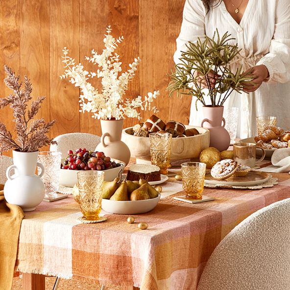 A woman stands behind a dining table filled with food, platters, glassware and vases. The table has a gingham tablecloth and cream coloured chairs, and the background is a wooden wall.