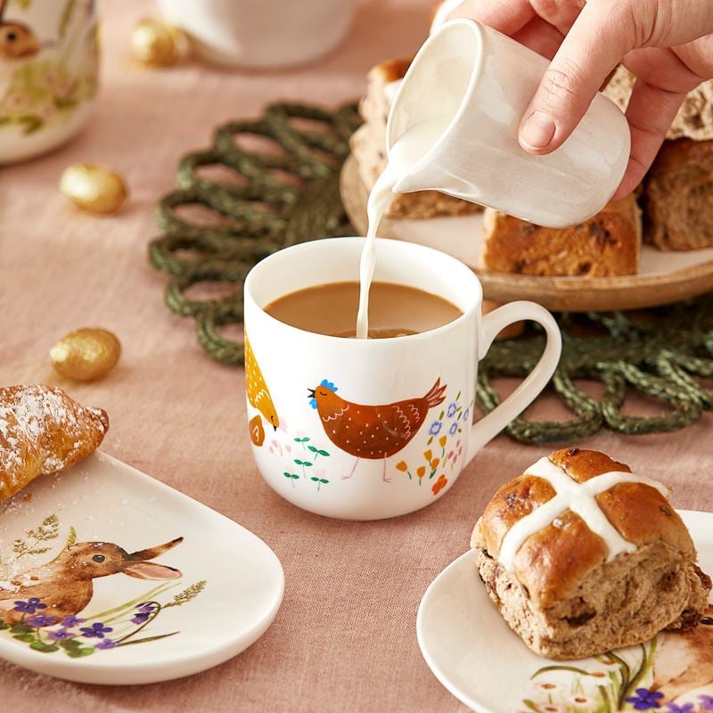 In the centre of the frame is a white mug with a hand pouring milk into the mug. Around the mug are baked treats on Easter themed plates and platters, and in the background are vases, Easter eggs and a plate of hot cross buns all on top of a pink linen tablecloth.