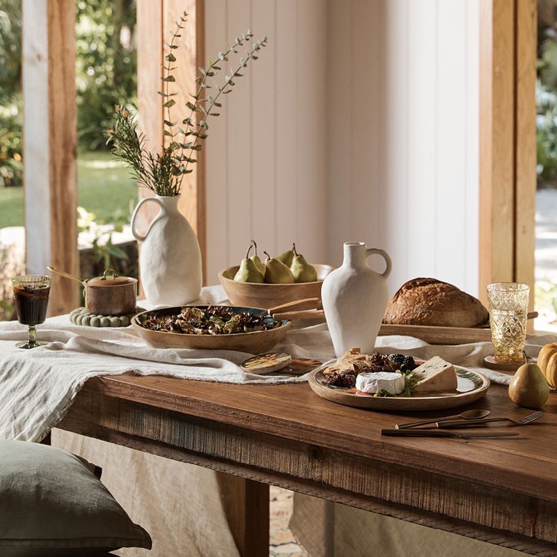 A timber table spread with an assortment of plates, platters, glasses and jugs all filled with fruits, cheeses and breads.