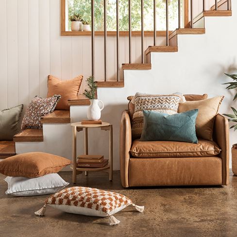 A tan leather armchair sits in front of a home staircase with an assortment of neutral tone cushions cascading down the bottom.