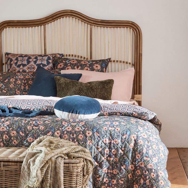 Front view of a bed with rattan bedhead in front of a white wall. The made bed has a floral bedspread with dark blue and green cushions.