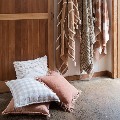 Four cushions sit stacked on the floor in front of a rustic timber door. To the right hang three throws in shades of brown and green.