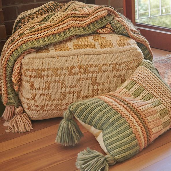 A close up picture of an ottoman, throw and cushion all with a chunky knitted pattern in shades of green, pink and tan.
