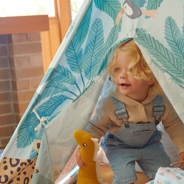 A child leans out of a tent, decorated with birds and fern leaves. The child is holding a toy, and the tent is in a living room.