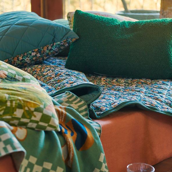 Close up shot of a mattress covered with a floral printed sleeping bag as well as green cushions and a blanket. The mattress has a rust coloured fitted sheet.