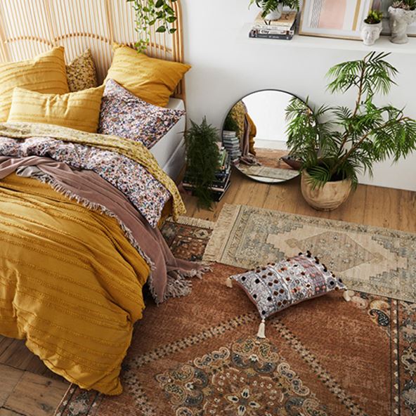 Bird’s eye view of bedroom with bed on left styled with mustard quilt cover and floral sheets, a selection of rugs and plants. 