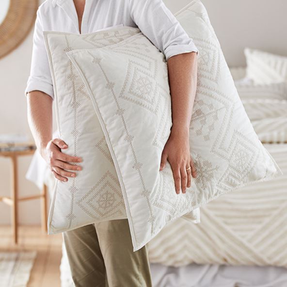 Person holding two neutral European pillows with detailed stitching and in the background is a bed with neutral bedlinen.  