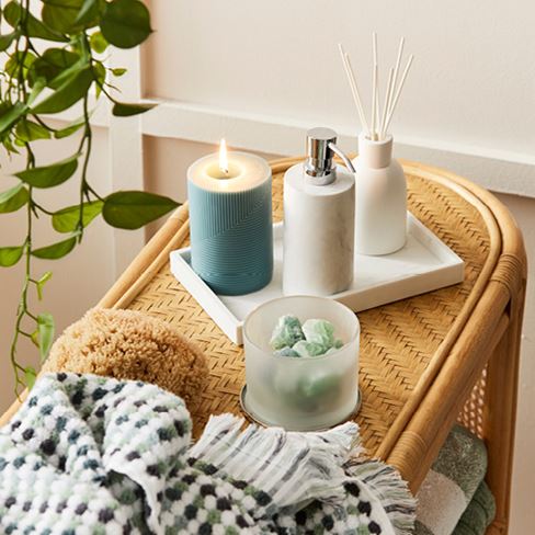 Rattan table styled with towel, candle, and selection of bathroom accessories on marble tray. 