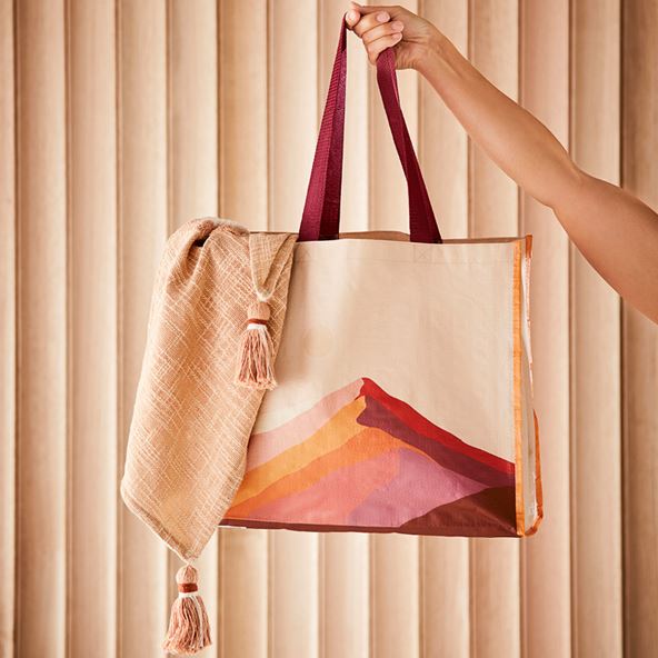 A hand holds up a tote bag with a mountain design. The background is a slatted wall.