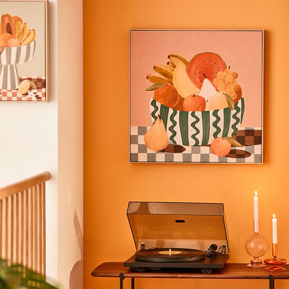 A record player and two candles sit on a wooden shelf, in front of a yellow wall. A piece of art with colourful fruit hangs on the wall.