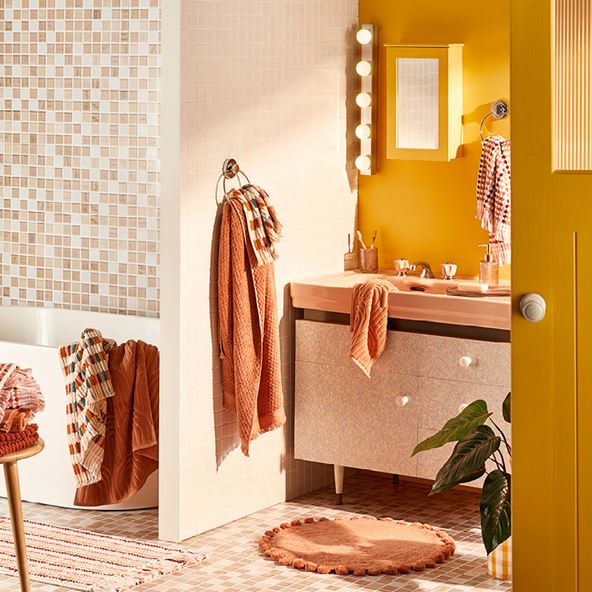 A bathroom scene with a bath to the left and vanity set up to the right. Towels and décor in shades of orange decorate the room and the tiles and paint are shades of yellow.