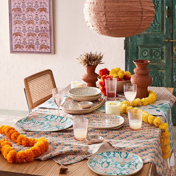 Natural wood table styled with warm blue, pink, yellow and amber hued runner, glass drinking glasses, stunning plates, vases, and more. 