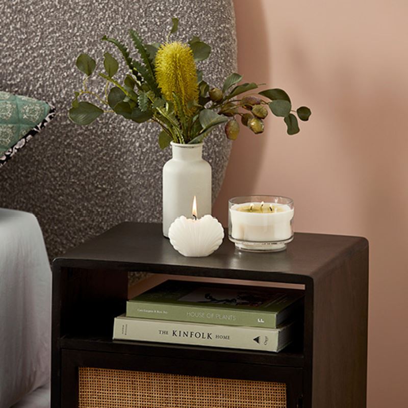 A black side table next to a grey bed, with a plant in a white vase and two small candles on top.