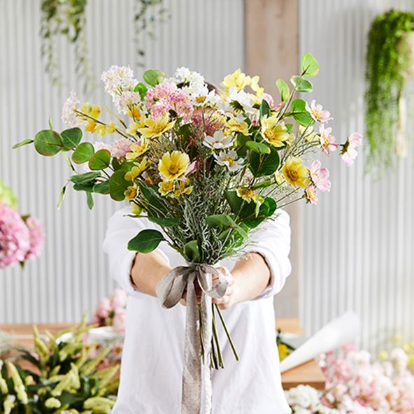Hands hold a colourful bunch of wildflowers, in shades of light pink and yellow amongst fresh green leaves.