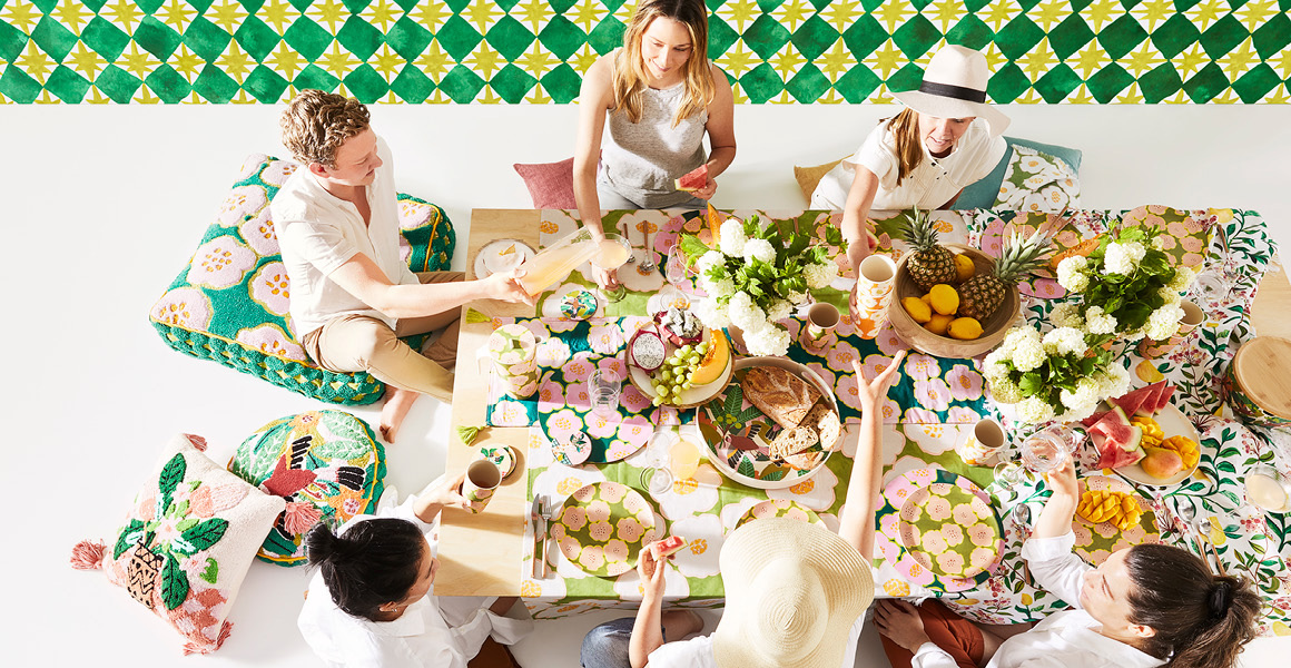 Friends gathered around a dining table enjoying an array of different food, the table is draped in a floral multicoloured tablecloth and various other tableware products from the Togetherness Collection. 