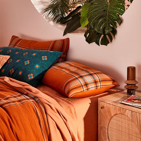 A close up image of a bed with printed cushions on shades of orange and blue, with a wooden bedside table to the right. Plants are hanging above the bed in the background.