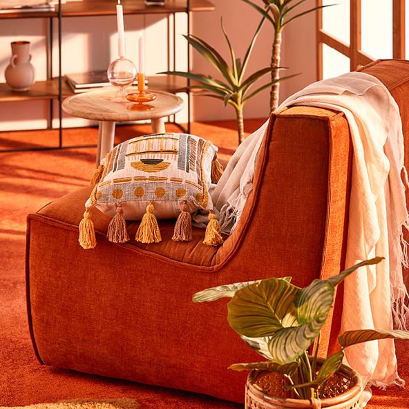 An orange chair viewed from the side, covered in textured cushions and throws. Plants and décor surround the chair, and the carpet is a similar shade of orange.