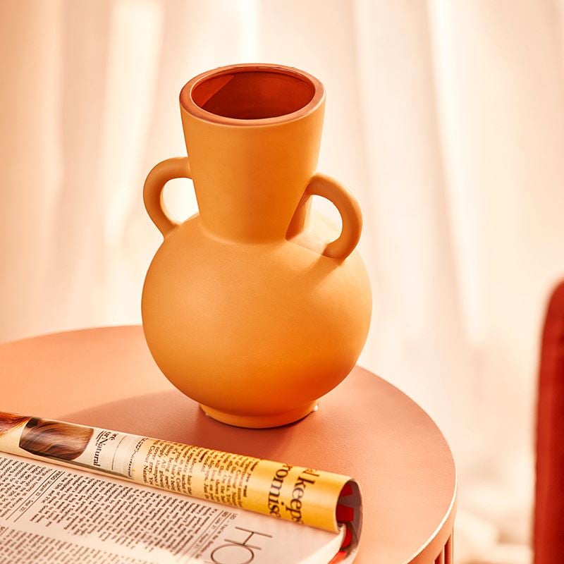 A close shot of a sculptural orange vase on a circular side table, with a magazine to the left corner of the frame.