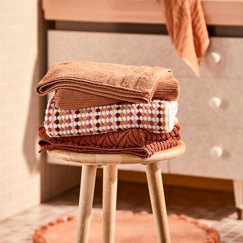 Textured towels in orange, clay and cream check are stacked on top of a wooden stool, with a bathroom vanity in the background.