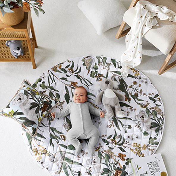Baby laying on a nursery play mat with botanical print, and matching accessories including plush koala toy and cushion. 