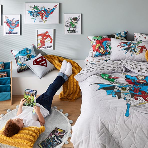 Bedroom with Justice League x Adairs Kids styled bed, accessories, wall art and a boy with his feet up on a ledge next to the bed reading a comic. 