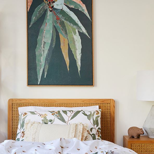 Arden Rattan Natural Bed with botanical bedlinen, neutral tufted cushion, and framed gum leaf wall art above the bed. 