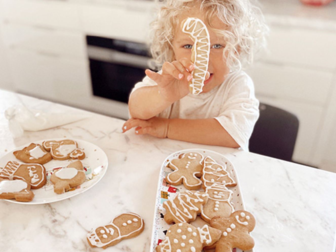 Toddler at kitchen bench holds a candy cane shaped ginger cookie in front of her face
