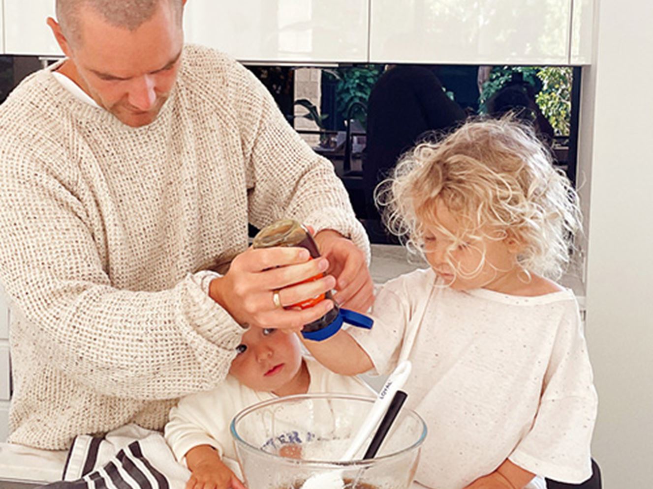 Father with two young children, baking Christmas gingerbread cookies, helps squeeze honey into a mixing bowl on a kitchen bench
