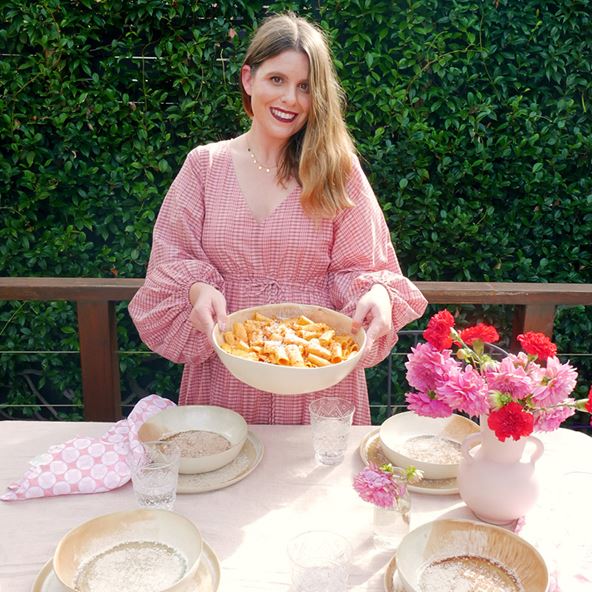 A woman in a pink and white check dress stands at an outdoor table, holding up a large bowl of pasta alla vodka. The table is set with bowls and plates, glasses and pink vase filled with pink flowers.